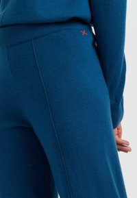 Blaue Strickhosen mit glatter Textur, ausgestattet mit einem Bund und einem kleinen roten Akzentstich. Nahaufnahme der Seitennähte.