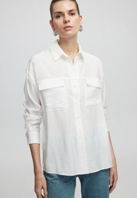 Camisa blanca de manga larga con botonadura frontal, dos bolsillos en el pecho y un corte relajado, hecha de tela ligera. Usada con denim azul.