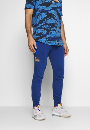 Blaue sportliche Jogginghosen mit orangefarbener Markenkennzeichnung und schmalem Schnitt. Getragen mit einem blauen Camouflage-Muster-Shirt und grauen Sneakers.