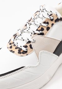 Zapatillas blancas con un upper de cuero texturizado, acentos de gamuza beige y una lengüeta de estampado de leopardo. Presentan una gruesa suela negra y cordones en contraste.