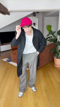 Fekete kabát, fehér póló, szürke kockás nadrág, ezüst cipő, rózsaszín kötött sapka. Beltéri környezet fa padlóval és zöld növénnyel.