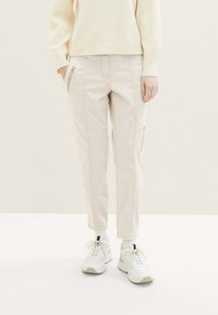 Personne portant un pantalon cargo slim beige clair avec des poches zippées et des baskets blanches, debout devant un fond crème uni.