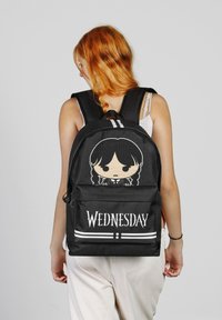 Mochila negra con un personaje de dibujos animados con trenzas y un atuendo formal. Gran texto que dice "MIÉRCOLES" en blanco. Correas ajustables.