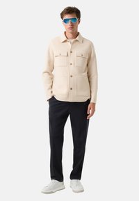 Jasno beżowa kurtka z wełny z dwiema kieszeniami na piersi, zapinana na guziki, w połączeniu z granatowymi spodniami i białymi sneakersami, oraz niebieskimi okularami przeciwsłonecznymi.