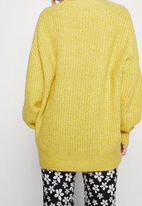 Osoba v přehnaně velkém žlutém pleteném svetru a černých kalhotách s bílým květinovým vzorem, viděná zezadu na jednobarevném pozadí.