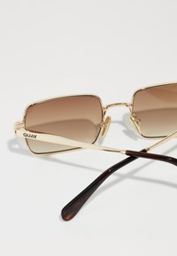 Lunettes de soleil rectangulaires avec monture en métal doré et verres dégradés marron. Les branches présentent un design écaille de tortue avec le logo QUAY.