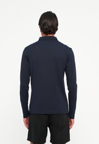 Mann med mørkt hår iført en marineblå langermet polo og svarte shorts, som står med ryggen mot en hvit bakgrunn.