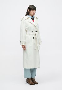 Lysebeige trenchcoat med dobbeltradet design, sorte knapper og bindebånd. Kombineret med lyseblå jeans og brune ankelstøvler.