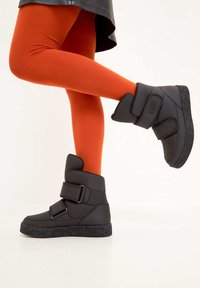 Botine negre până la gleznă cu două curele late cu Velcro, talpă din cauciuc texturat și partea superioară din material sintetic neted, purtate cu colanți portocalii.