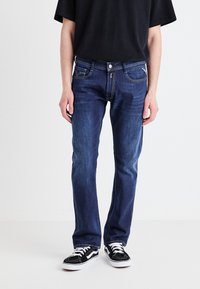 Mörkblå denimjeans med rak passform, framfickor och kontrasterande sömmar. Kombinerade med svarta sneakers med vita accenter.