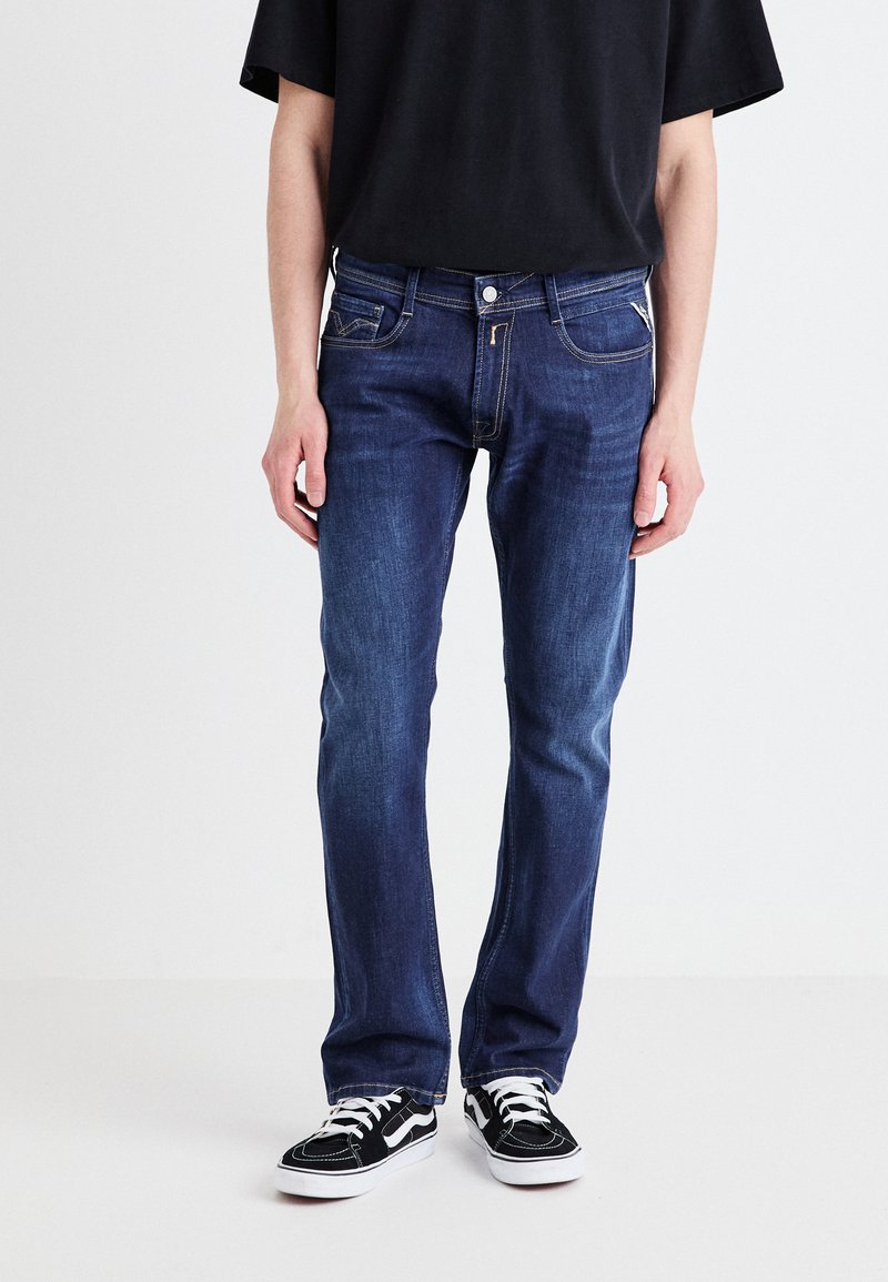 Mörkblå denimjeans med rak passform, framfickor och kontrasterande sömmar. Kombinerade med svarta sneakers med vita accenter.