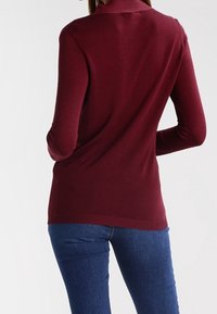 Burgundský lehký svetr s vysokým límcem a dlouhými rukávy. Přiléhavý střih s žebrovaným lemem, kombinovaný s modrými džíny.