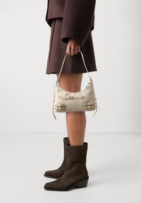 Beige leather handbag with two buckle accents, soft rounded design, and short handle. Model wears a dark brown coat and boots.