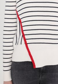 Pull en maille blanche à rayures bleu marine avec un détail latéral rouge. Il présente une texture côtelée et un ourlet côtelé, avec des manches longues et une coupe décontractée.