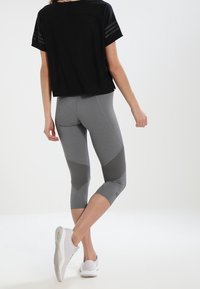 Žena v černém topu s krátkým rukávem, šedých legínách ke kotníkům s tmavšími panely a bílých sportovních botách stojící na čistém bílém pozadí.