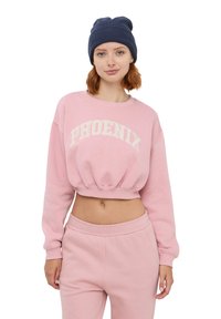 Felpa corta rosa con la scritta "PHOENIX" in bianco con testo arcuato, tessuto in pile testurizzato. Abbinata a pantaloni della tuta rosa coordinati, berretto in maglia morbida.