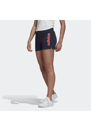 Person, die marineblaue Adidas-Shorts mit pinkem Logo und weißen Turnschuhen trägt, vor hellem Hintergrund steht.
