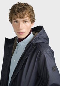 Námořnická vodotěsná bunda s kapucí, s hřejivou podšívkou a patentkami. Nositelná přes světle modrou košili, s logem na rukávu.