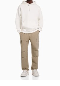 Bíla mikina s kapucí a stahovací šňůrkou, velká přední kapsa. Béžové cargo kalhoty s bočními kapsami a volnějším střihem. Bílé tenisky doplňují vzhled.