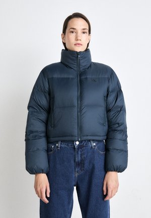 Pufferjacke in donkerblauw, met een hoge nek, ritssluiting en oversized mouwen. Gekleed met high-waisted denim jeans.