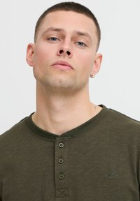 Olivgrünes Henley-Shirt mit drei Knöpfen und geripptem Kragen. Verfügt über ein kleines gesticktes Logo auf der Brust und einen weichen, strukturierten Stoff.