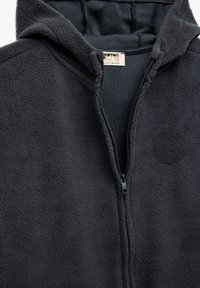 Sort fleece zip-up hættejakke med en blød tekstur, der har en frontlynlås og en foret hætte. Mærket viser "Keton Kids, 5-9Y."