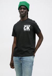 Čierne bavlnené tričko s bielym nápisom "CALVIN KLEIN CK Established 1968." Kombinované so svetlomodrými džínsami a zelenou pletenou čiapkou.