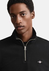 Zwarte pullover met ritssluiting gemaakt van gladde stof, met een hoge kraag en een metalen rits. Opvallend geborduurd logo op de linkerborst.