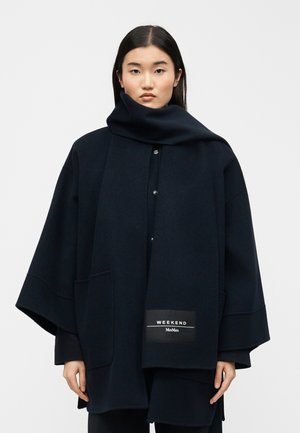 Frau trägt einen dunklen, übergroßen Mantel und einen passenden langen Schal mit dem Etikett "WEEKEND MaxMara" auf dem Schal vor einem einfarbigen Hintergrund.