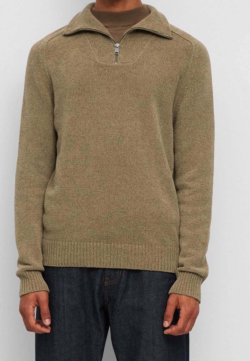 Pull en tricot kaki avec un col zippé à moitié, des poignets et un ourlet côtelés, un tissu texturé et une coupe décontractée, associé à un jean en denim foncé.
