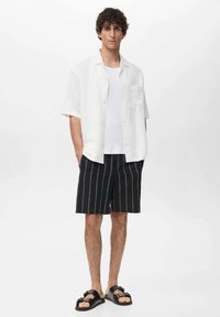 Witte kortebloemige blouse over een witte tanktop, gecombineerd met zwarte gestreepte shorts en zwarte sandalen. Eenvoudig ontwerp, relaxte pasvorm.