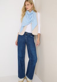 Una bufanda azul clara se coloca sobre una camisa blanca de manga larga, combinada con un chaleco beige y jeans de pierna ancha de color azul oscuro. Zapatillas beige completan el look.