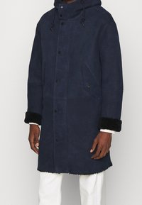 Manteau long bleu marine en daim doux avec capuche, doté de poignets noirs duveteux et de poches avant à fermeture pression.