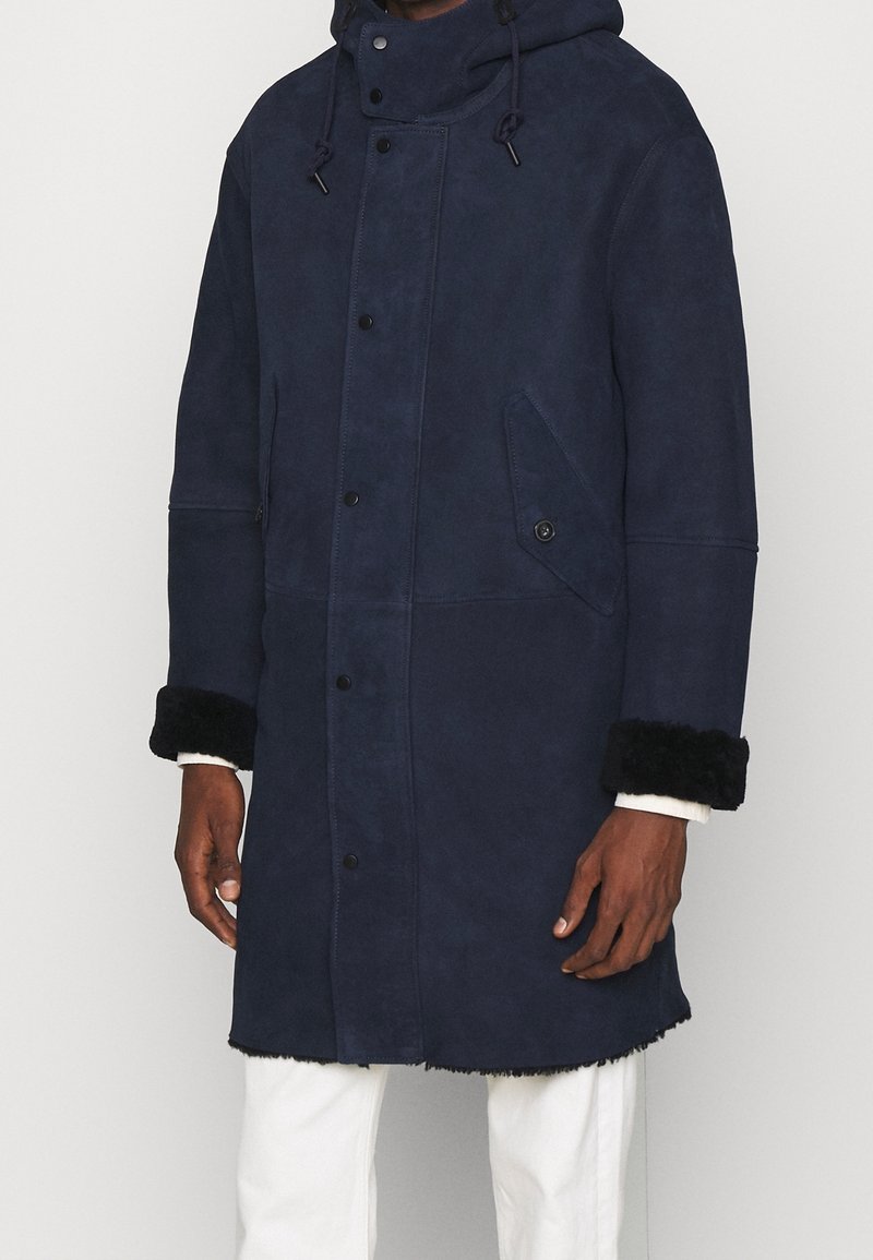 Manteau long bleu marine en daim doux avec capuche, doté de poignets noirs duveteux et de poches avant à fermeture pression.