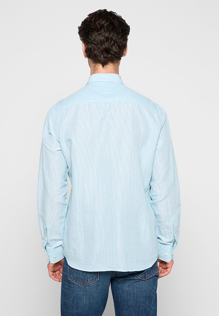 Mann mit dunklem, lockigem Haar, der ein hellblaues, langärmliges Hemd und eine Jeans trägt, mit dem Rücken zur Kamera.