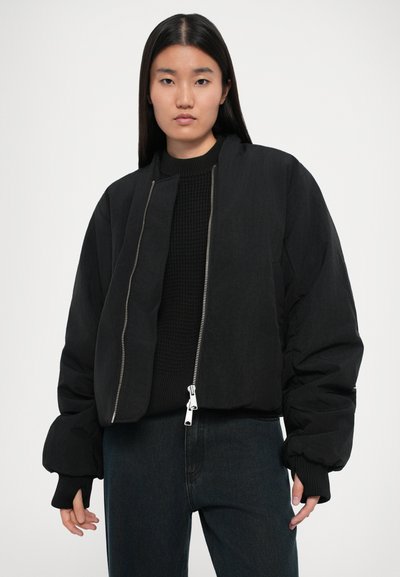 Chaqueta bomber negra corta con una cremallera plateada brillante, puños elásticos y un suéter de punto negro texturizado debajo. Combinada con denim oscuro.