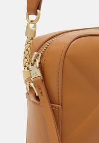 Brązowa torebka na ramię z jasnobrązowej skóry z elementami w kolorze złotym, z zamkiem błyskawicznym i ozdobnymi splotami łańcucha. Gładka faktura i wyraźnie zarysowany kształt.