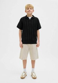Polo shirt noir à manches courtes en maille avec un col en V, présentant des rayures verticales. Associé à un short beige au genou et des baskets beiges avec des accents blancs.