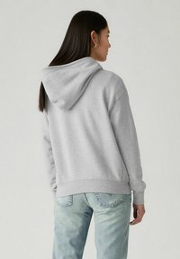 Felpa grigia con cappuccio e vestibilità ampia, con polsini e orlo a costine. Caratterizzata da un cappuccio con cordino e da un tessuto morbido e strutturato. Indossata con jeans azzurri chiari.