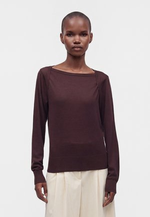 Donna con capelli corti indossa una maglia marrone scuro a maniche lunghe e pantaloni larghi beige chiaro, in piedi davanti a uno sfondo chiaro e uniforme.