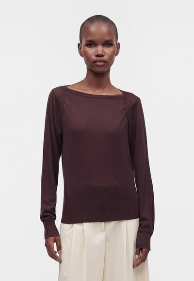 Femme aux cheveux courts porte un haut à manches longues marron foncé et un pantalon large beige clair, debout devant un fond clair uni.