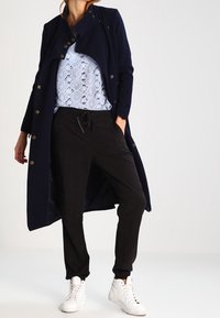 Femme portant un long manteau marine, un haut à motifs bleus, un pantalon noir à cordon de serrage et des baskets montantes blanches sur fond uni.