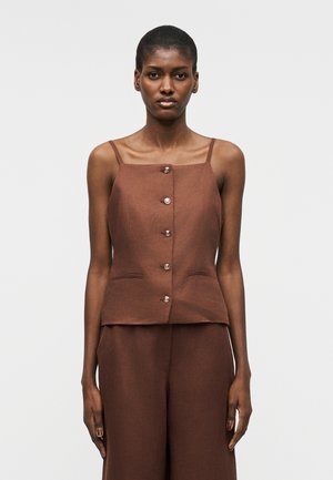 Frau trägt ärmelloses braunes Oberteil mit dünnen Trägern und passenden, weit geschnittenen Hosen, steht vor einem schlichten hellen Hintergrund.