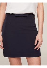 Woman wearing a navy blue skirt with a small bow detail at the waistband and a white tucked-in top, standing with hands relaxed by sides.
