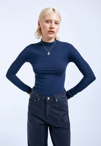 Dunkelblaues Langarmshirt mit slim fit, hohem Kragen und gerafften Ärmeln, kombiniert mit dunklen, hochgeschnittenen Jeans mit Fronttaschen.
