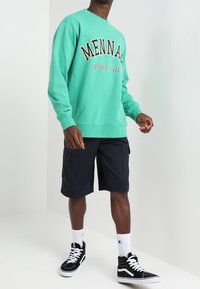Felpa verde menta con testo "MENNAH SPORTS WEA", abbinata a pantaloni cargo blu scuro, sneakers alte nere e calzini bianchi a righe.