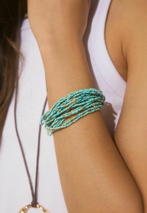 Arm wearing multiple strands of turquoise and gold beaded bracelet over a white sleeveless top.