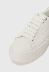 Vit lädersneaker med platta vita snören, rund tå och texturerad gummisula. Har små perforeringar för andningsförmåga.