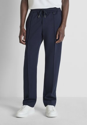 Mann trägt marineblaue gerade Hose mit Tunnelzug an der Taille und weiße Sneakers, steht vor einem schlichten hellen Hintergrund.
