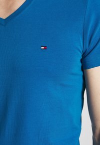 Blaues Baumwoll-V-Ausschnitt-T-Shirt mit einem kleinen gestickten Logo in Rot, Weiß und Schwarz auf der linken Brust. Glatte Textur, kurze Ärmel.